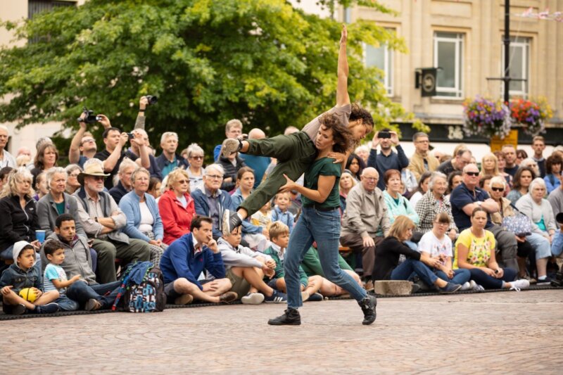 One dancer lifts another, balanced on her side, in front of a large outdoor audience in Newbury market place.