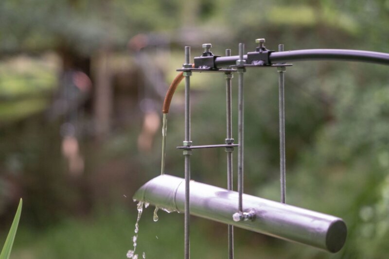 A close up of a metal tube gently pouring water, set against a green lush pondscape.