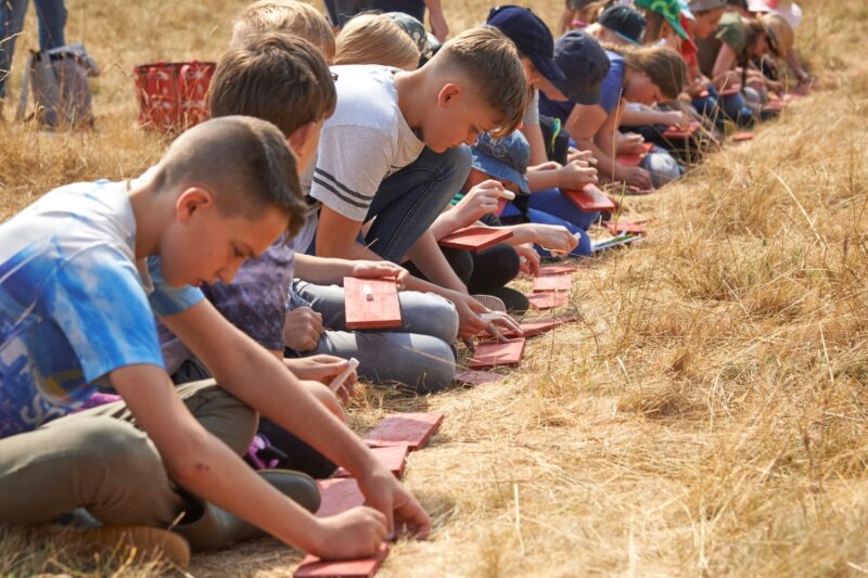 Children sit in a row in a grassland landscape, writing or drawing in books.