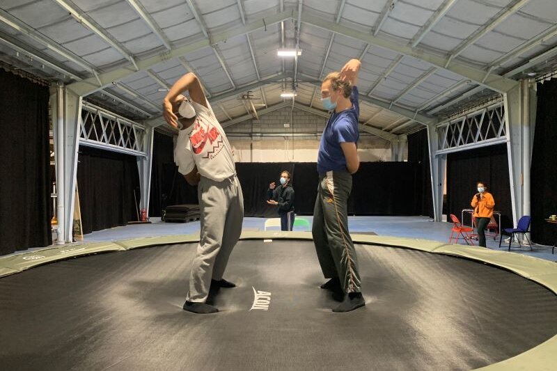 Two dancers face each other on a trampoline, leaning backwards from the knees