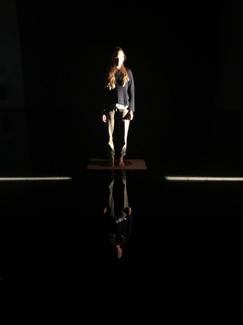 A woman with long red hair, wearing a black top and khaki trousers stands on a platform in a pitch black room with one stripe of white light running along the floor behind her. Her reflection is mirrored in the shiny floor beneath her.