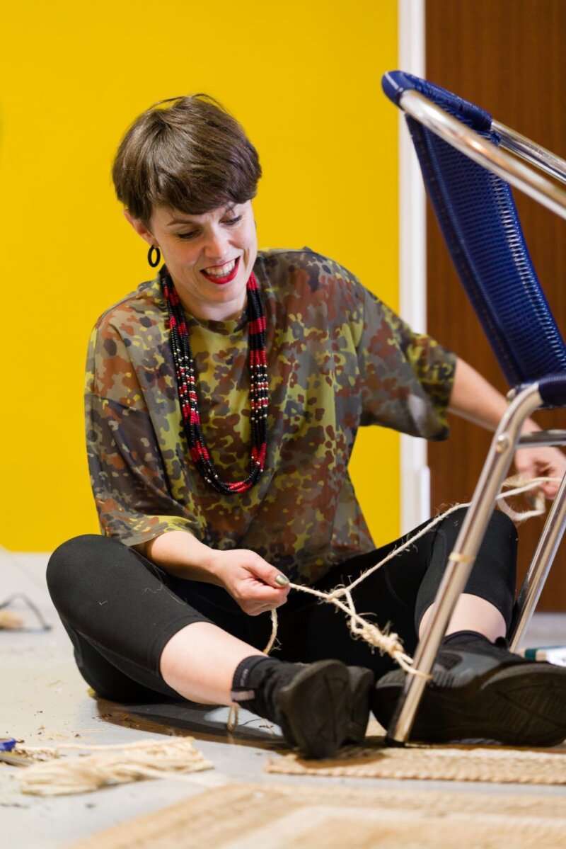 A woman seated on the ground is tying string around the legs of a folding chair.