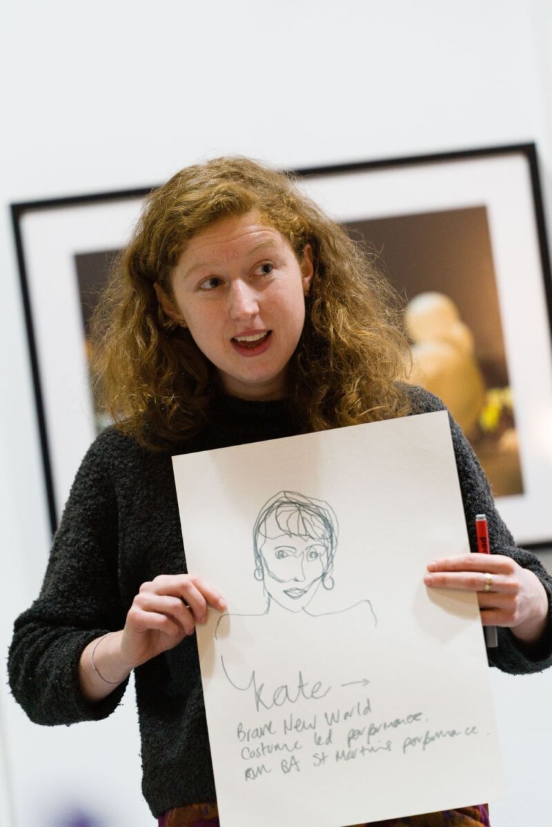 A woman with thick wavy long red hair holds up a drawing of a woman with a bob. Handwriting on the drawing says 'Kate' and there is more description written underneath.